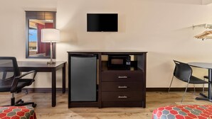 Quarto de hotel com escrivaninha, frigorífico, micro-ondas, televisão, cadeira e cama com colcha estampada.