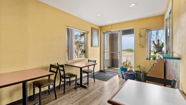 Quarto pequeno e acolhedor, com piso de madeira, duas mesas e cadeiras. Possui duas janelas com cortinas, uma porta que dá para o exterior e uma pequena varanda com plantas em vasos.