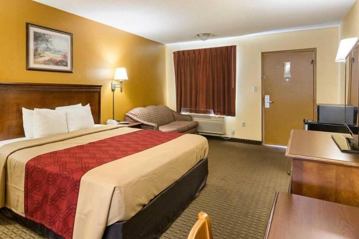 Habitación de hotel con cama, escritorio, silla, lámpara, ventana con cortinas y puerta.