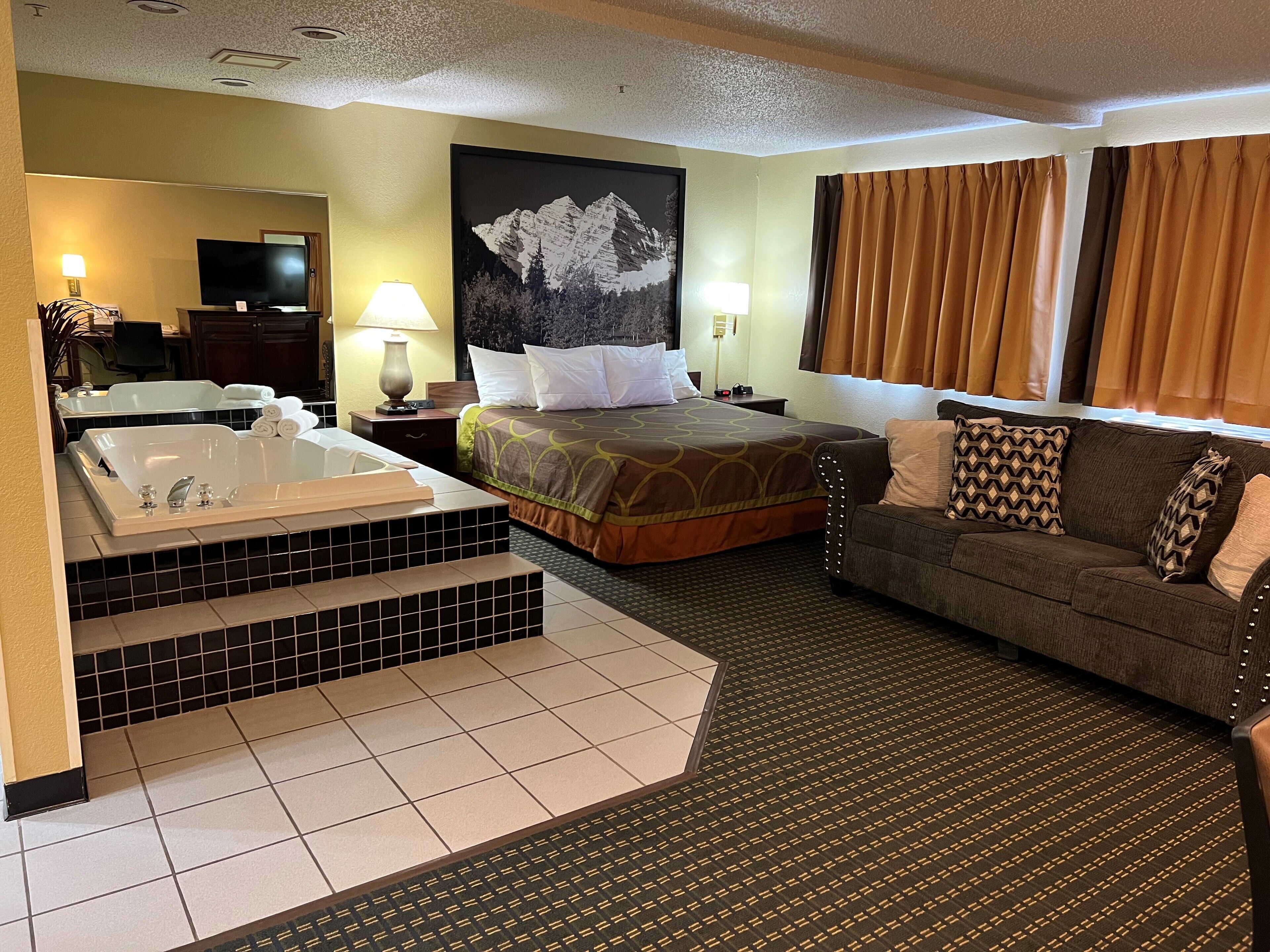 A hotel room with a large bed, a sofa, a bathtub, and a mountain painting on the wall.