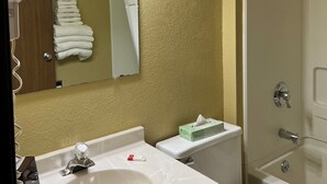 Une salle de bain avec un lavabo, des toilettes et une douche. Des serviettes sont pliées et empilées sur un porte-serviettes au mur, il y a un miroir et un luminaire au-dessus du lavabo.