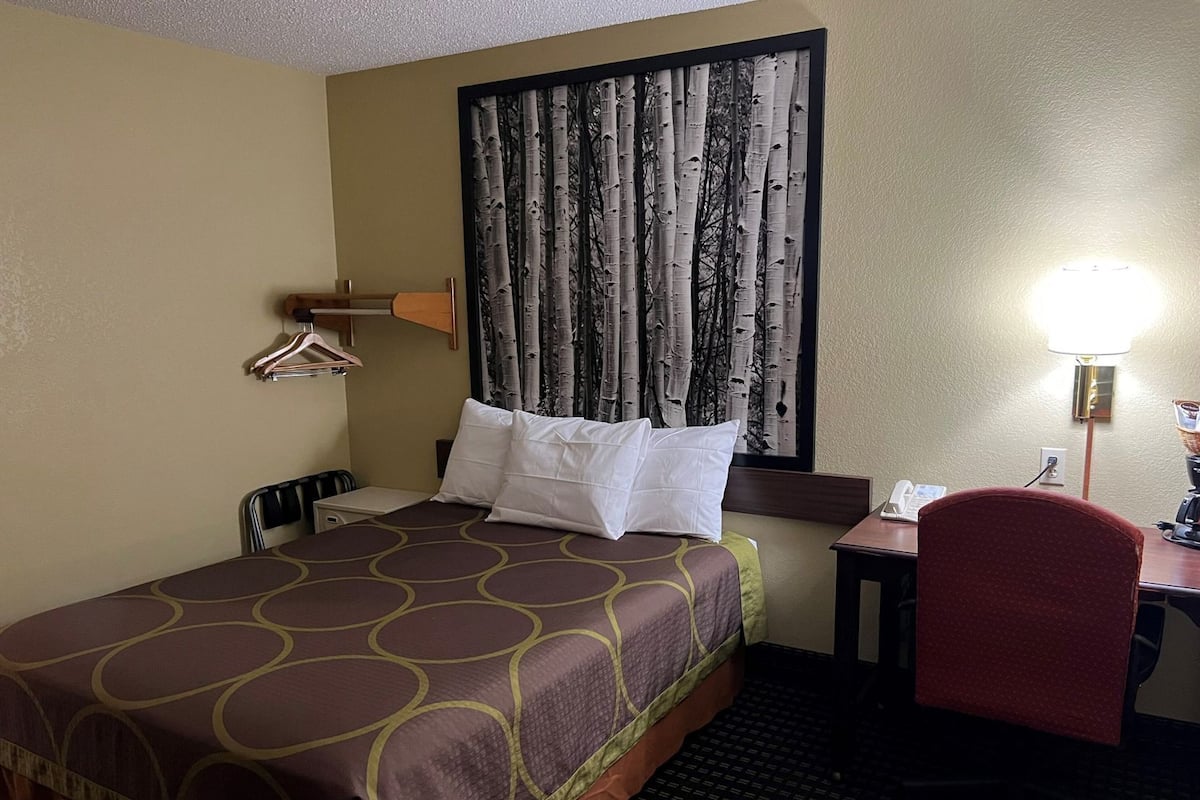 Une chambre d’hôtel avec un lit, un bureau, une chaise, une lampe et une fenêtre avec des rideaux.