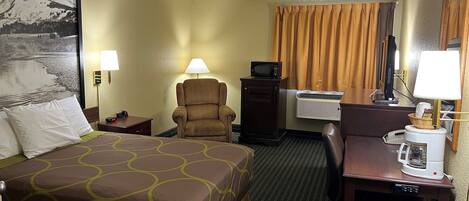 A hotel room with a bed, a desk with a computer, a chair, a desk lamp, a microwave, an air conditioner, and a large framed picture on the wall.