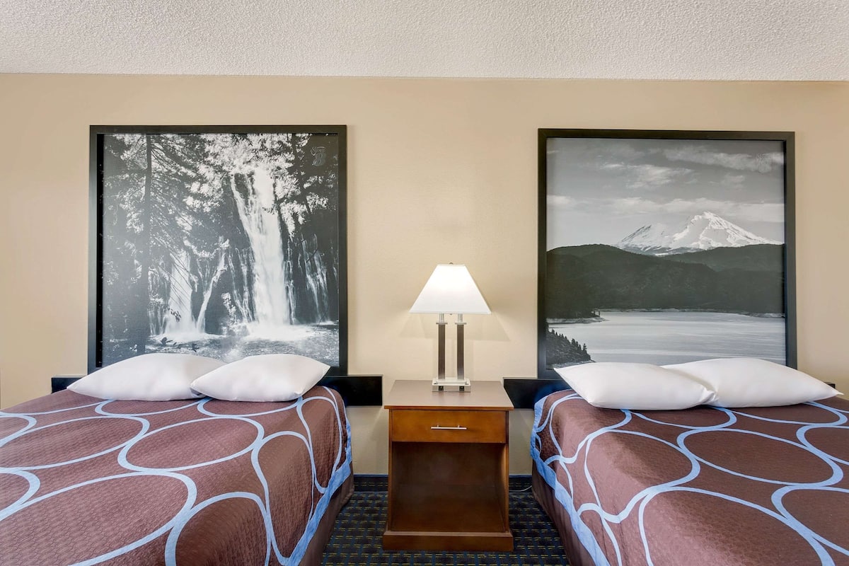 A hotel room with two beds, a nightstand with a lamp, and two large framed pictures on the walls.