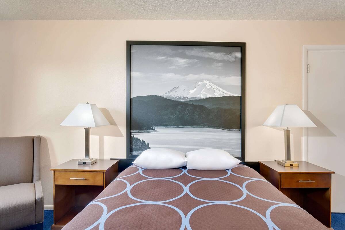 A hotel room with a bed, two bedside tables with lamps, a chair, and a large framed picture of a mountain.
