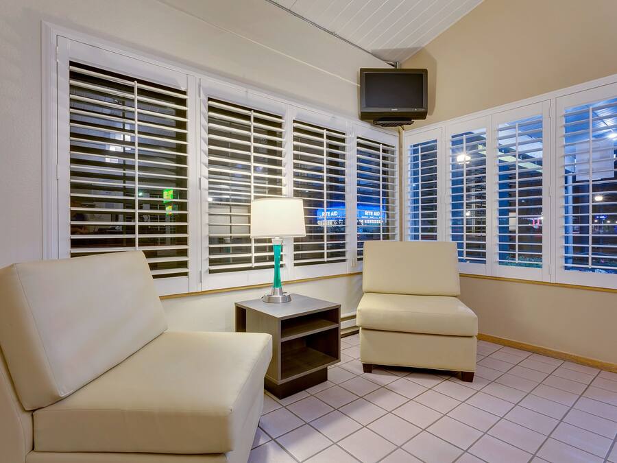 A room with two beige sofas, a small side table with a lamp, a television mounted on the wall, and large windows with blinds.
