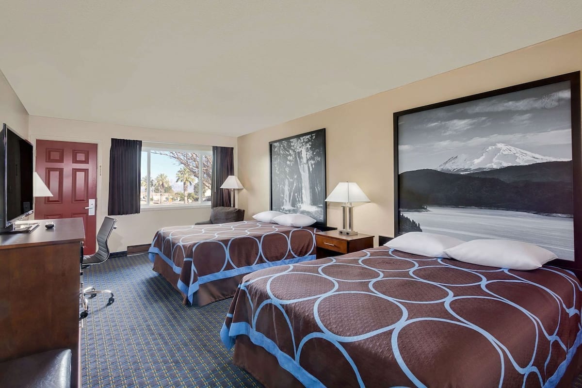 A hotel room with two beds, a desk, a chair, a TV, and a large window with a mountain view.