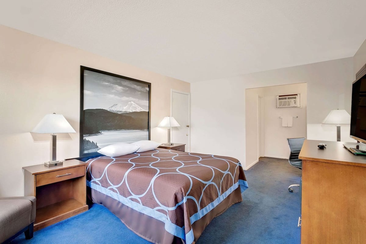 A hotel room with a bed, two bedside tables with lamps, a desk with a chair, a large window with a view of mountains, and a door.