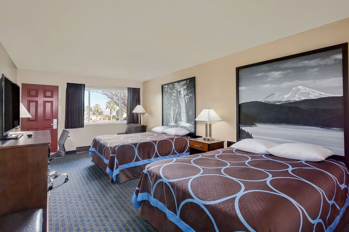 A hotel room with two beds, a desk, a TV, and a large window with a mountain view.