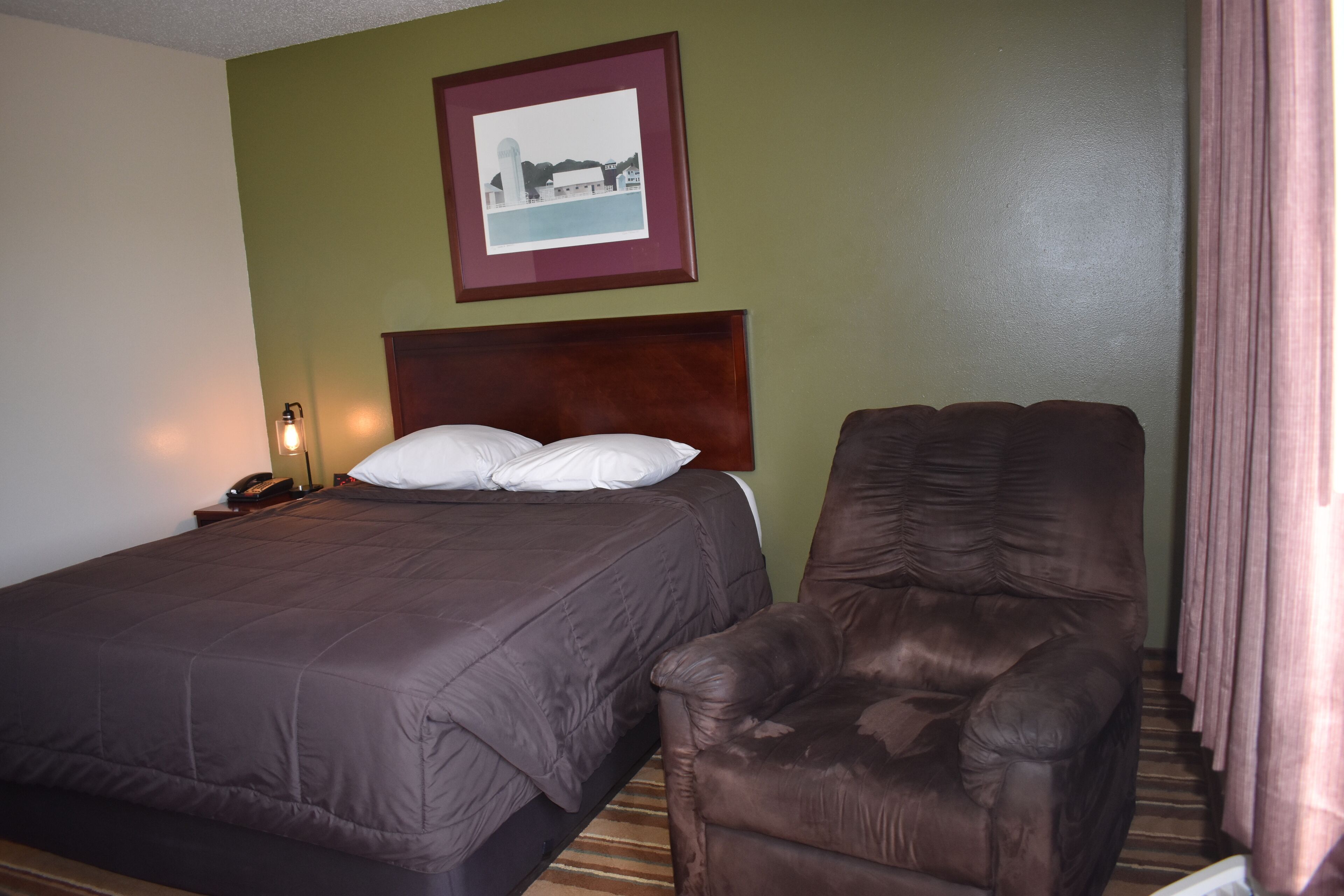 A hotel room with a bed, a chair, a nightstand with a lamp, and a framed picture on the wall.