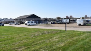 A parking area with a large building, a white fence, and several vehicles including a white RV and a white truck. - Heartland Hotel and Suites (Rock Valley)