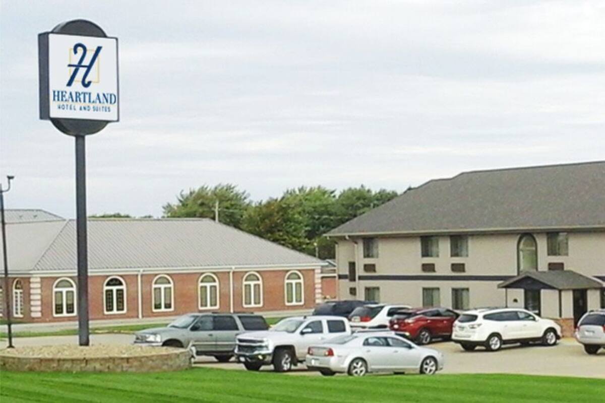 A Heartland Hotel and Suites with a parking lot filled with cars, a brick building, and a large, flat-roofed structure.