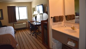 A hotel room with a bed, desk, chair, and a large mirror above the sink. - Heartland Hotel and Suites (Rock Valley)