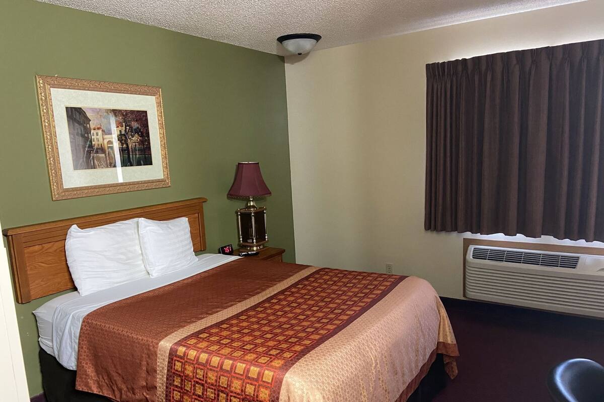 A hotel room with a bed, a nightstand with a lamp, a framed picture on the wall, a window with curtains, and an air conditioning unit.