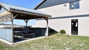 An outdoor barbecue area with a covered picnic table and grill, adjacent to a building with a door and windows. - Heartland Hotel and Suites (Rock Valley)