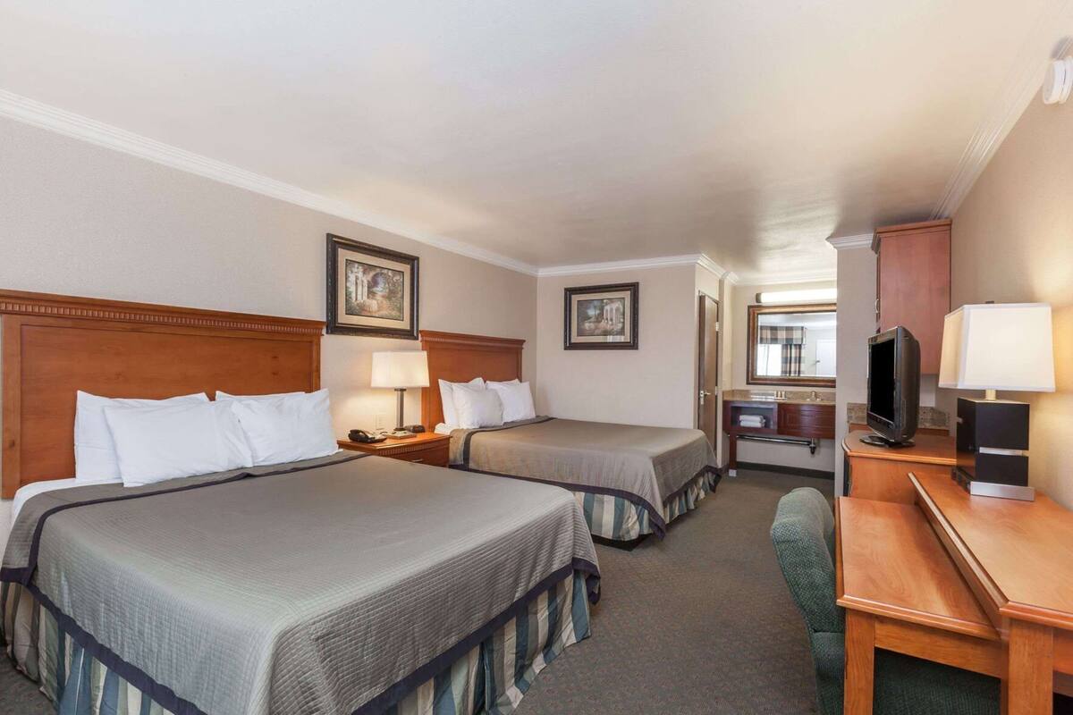 Habitación de hotel con dos camas, un escritorio, un televisor y un baño visible al fondo.