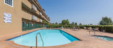 Una piscina con escalera metálica, rodeada por un solárium con baldosas y un edificio con varios balcones y un cartel.