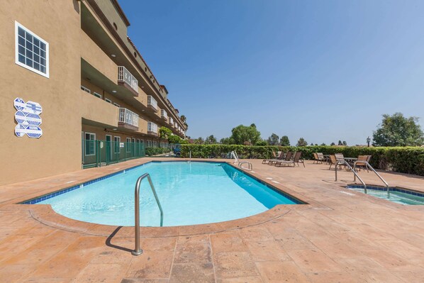 Una piscina con escalera metálica, rodeada por un solárium con baldosas y un edificio con varios balcones y un cartel.