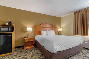 A hotel room with a bed, a desk with a microwave, a nightstand with a lamp, and a window with curtains.