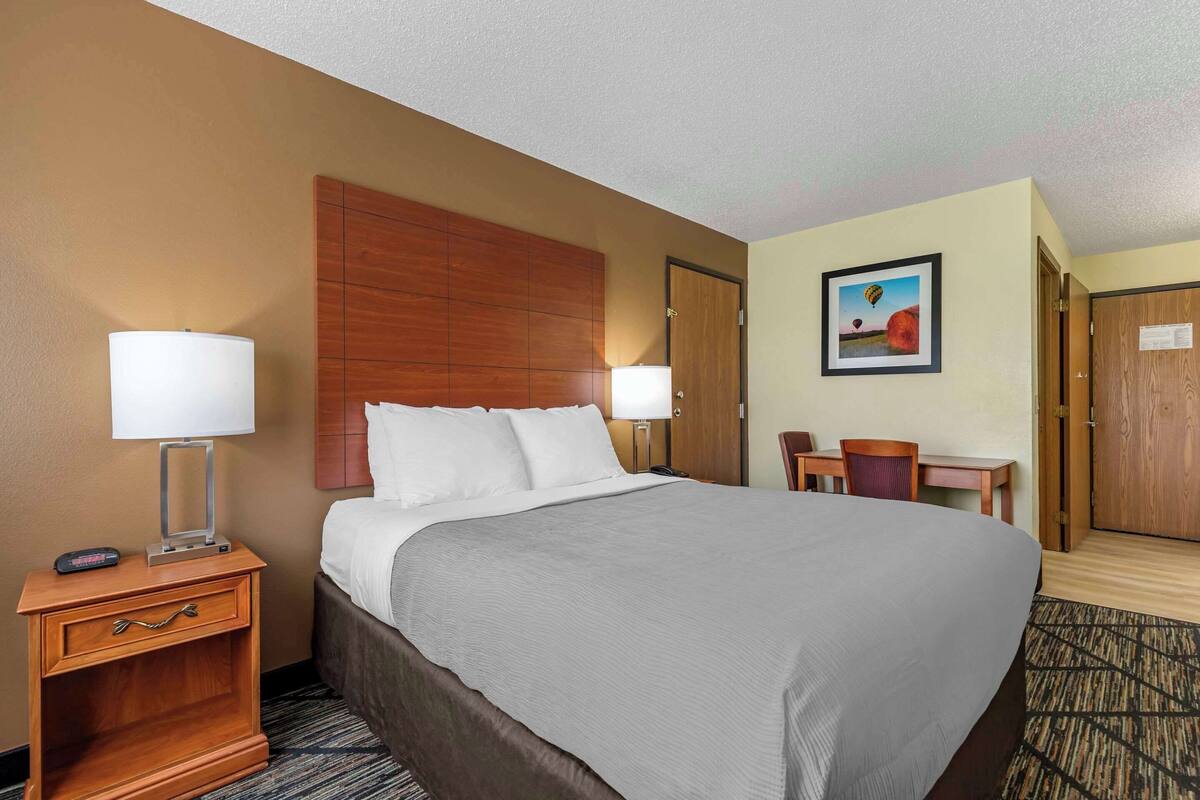 A hotel room with a large bed, two bedside lamps, a wooden headboard, a desk with a chair, and a painting on the wall.