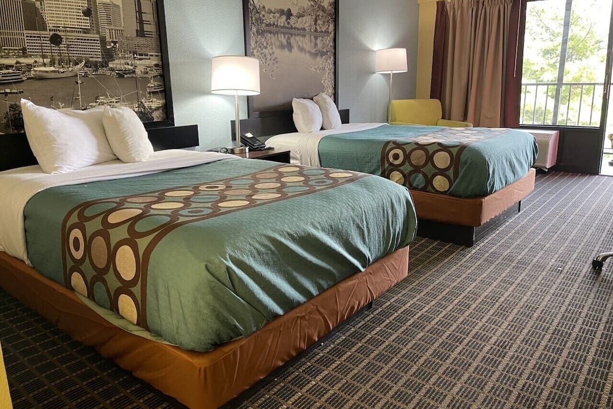 A hotel room with two beds, a large window with curtains, a cityscape view, and a patterned carpet.