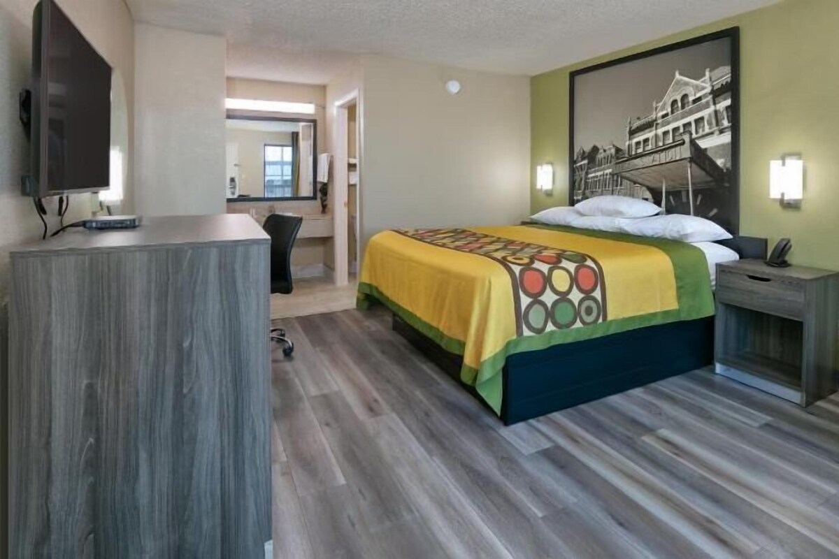 A hotel room with a bed, a desk, a chair, a TV, and a wall-mounted light.