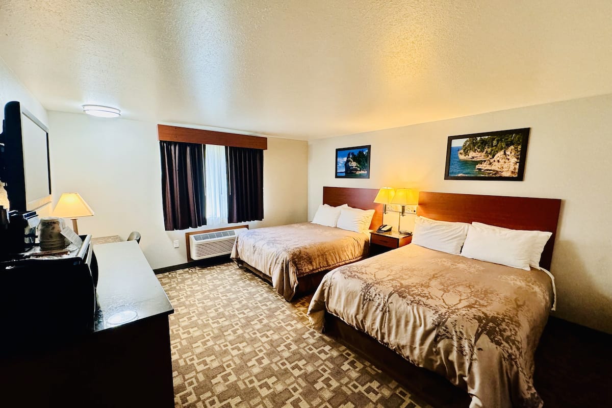 Une chambre d’hôtel avec deux lits, un bureau, une lampe, une fenêtre avec des rideaux, un climatiseur fixé au mur et deux tableaux encadrés.