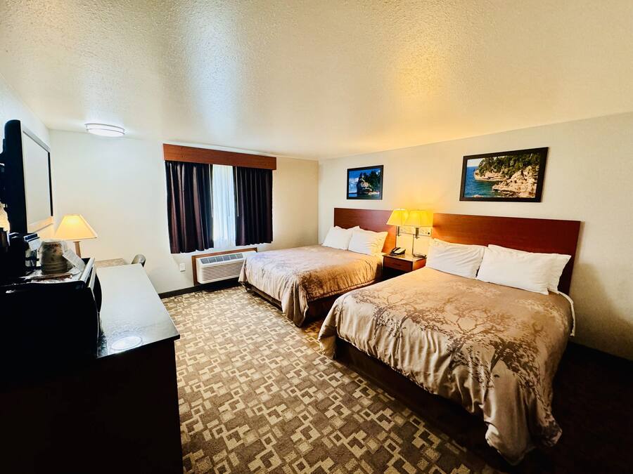 Une chambre d’hôtel avec deux lits, un bureau, une lampe, une fenêtre avec des rideaux, un climatiseur fixé au mur et deux tableaux encadrés.