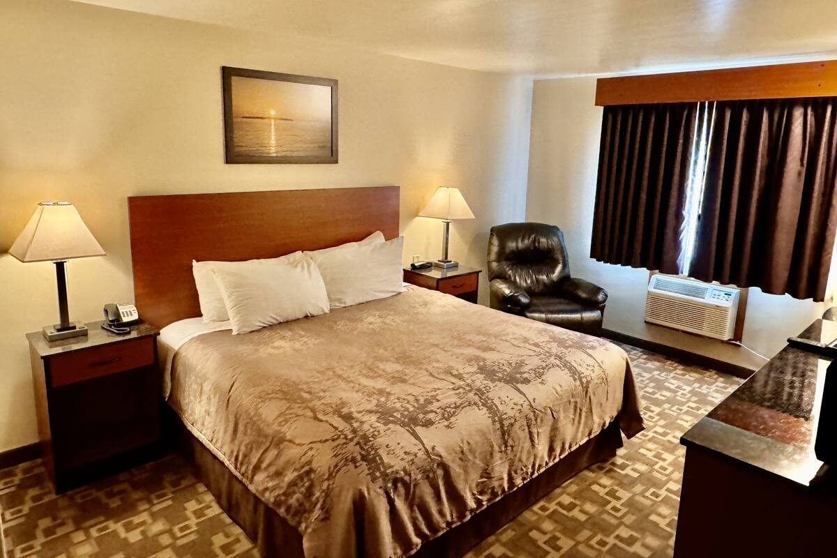 Une chambre d’hôtel avec un grand lit, deux lampes de chevet, un fauteuil en cuir, un radiateur et une fenêtre avec des rideaux.