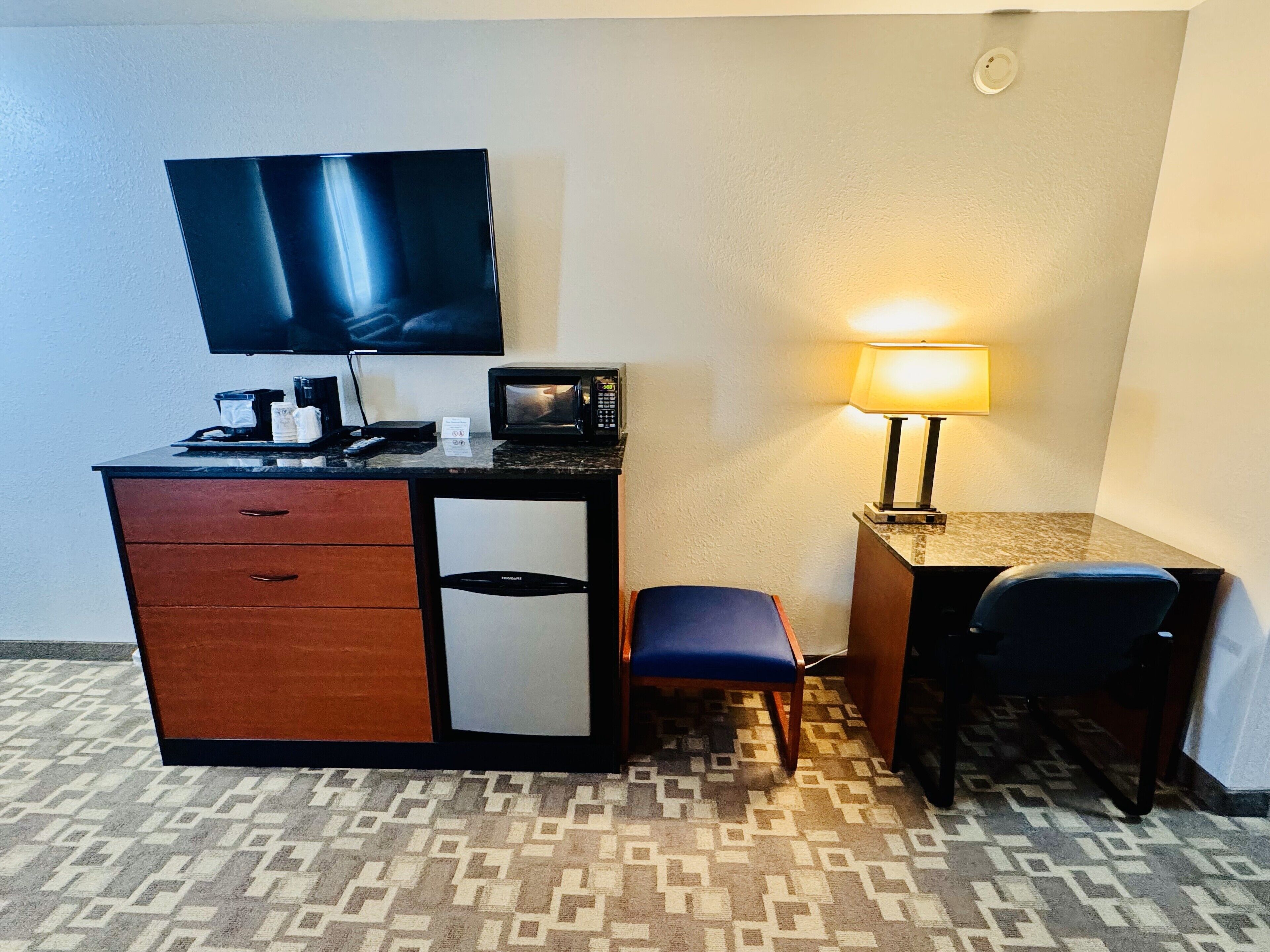 A hotel room with a desk, a television, a microwave, a refrigerator, a chair, and a lamp.