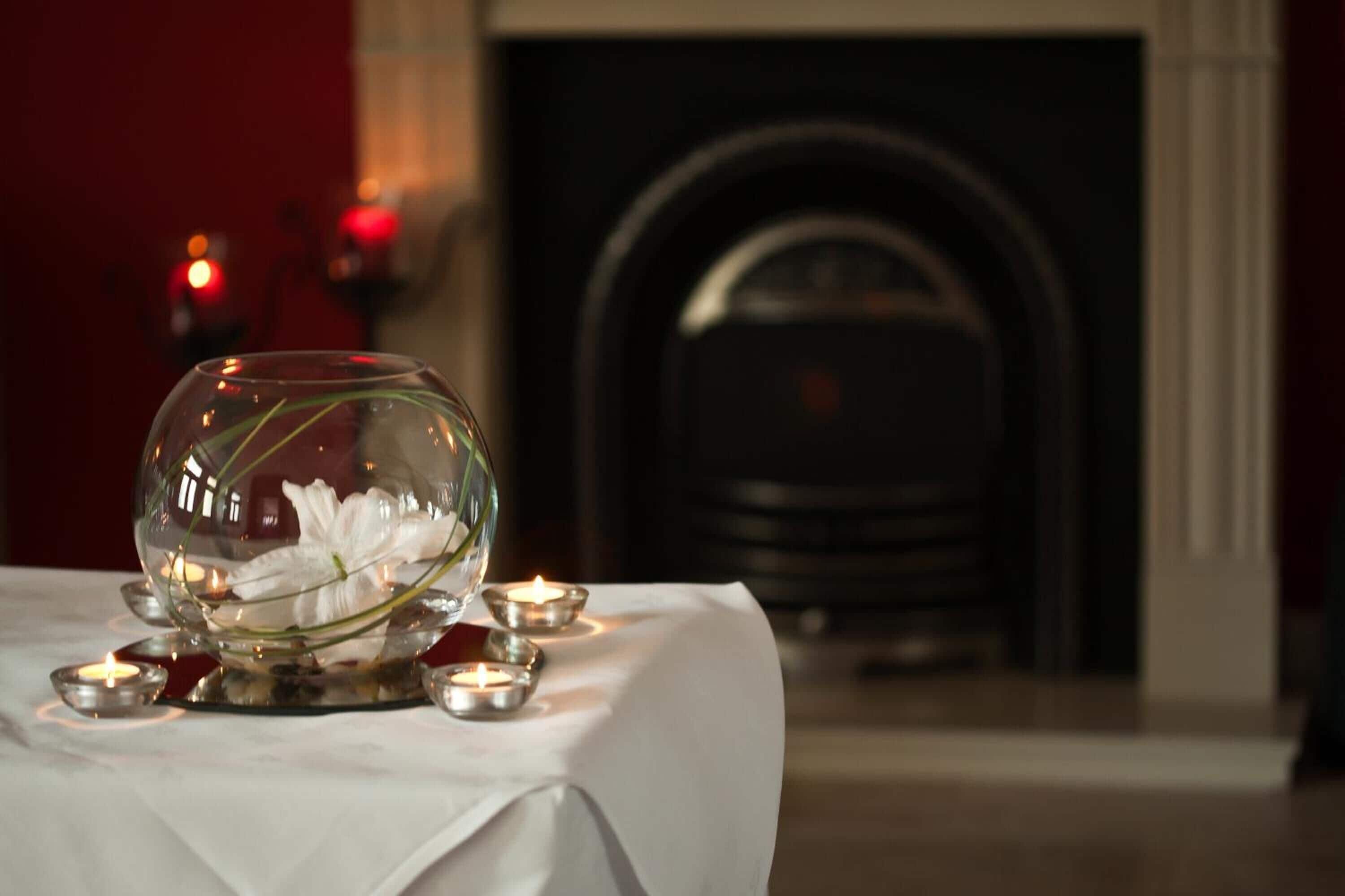 Una mesa de comedor con un bol de vidrio que contiene flores blancas, velas y una chimenea al fondo.