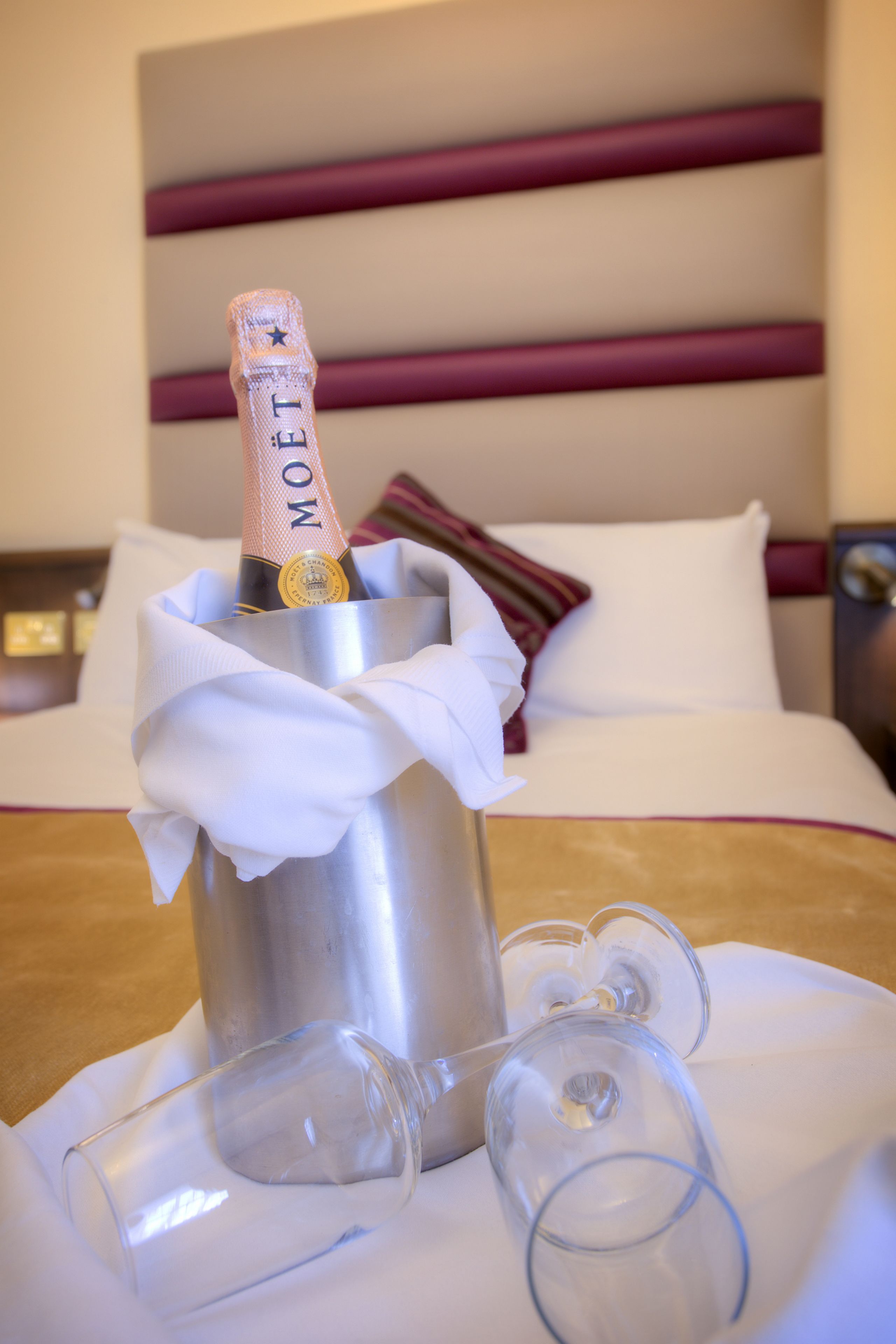 A minibar with a bottle of Moët champagne, two empty wine glasses, and a white cloth napkin on a bed with white bedding and a striped pillow.