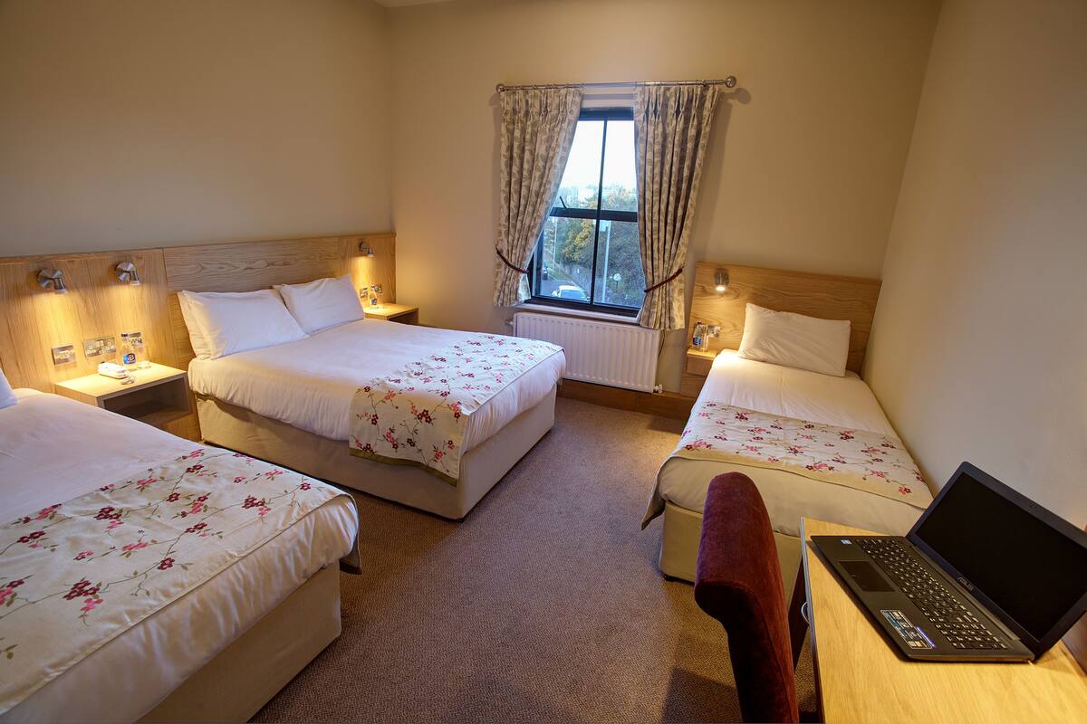 A hotel room with three beds, a laptop on a desk, and a window with curtains.