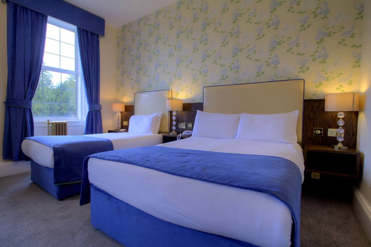 A hotel room with two beds, a window with curtains, floral wallpaper, and bedside lamps.