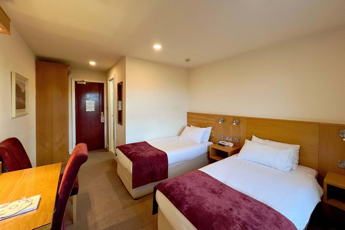 A hotel room with two beds, a wooden headboard, a desk with a chair, and a door leading to another room.