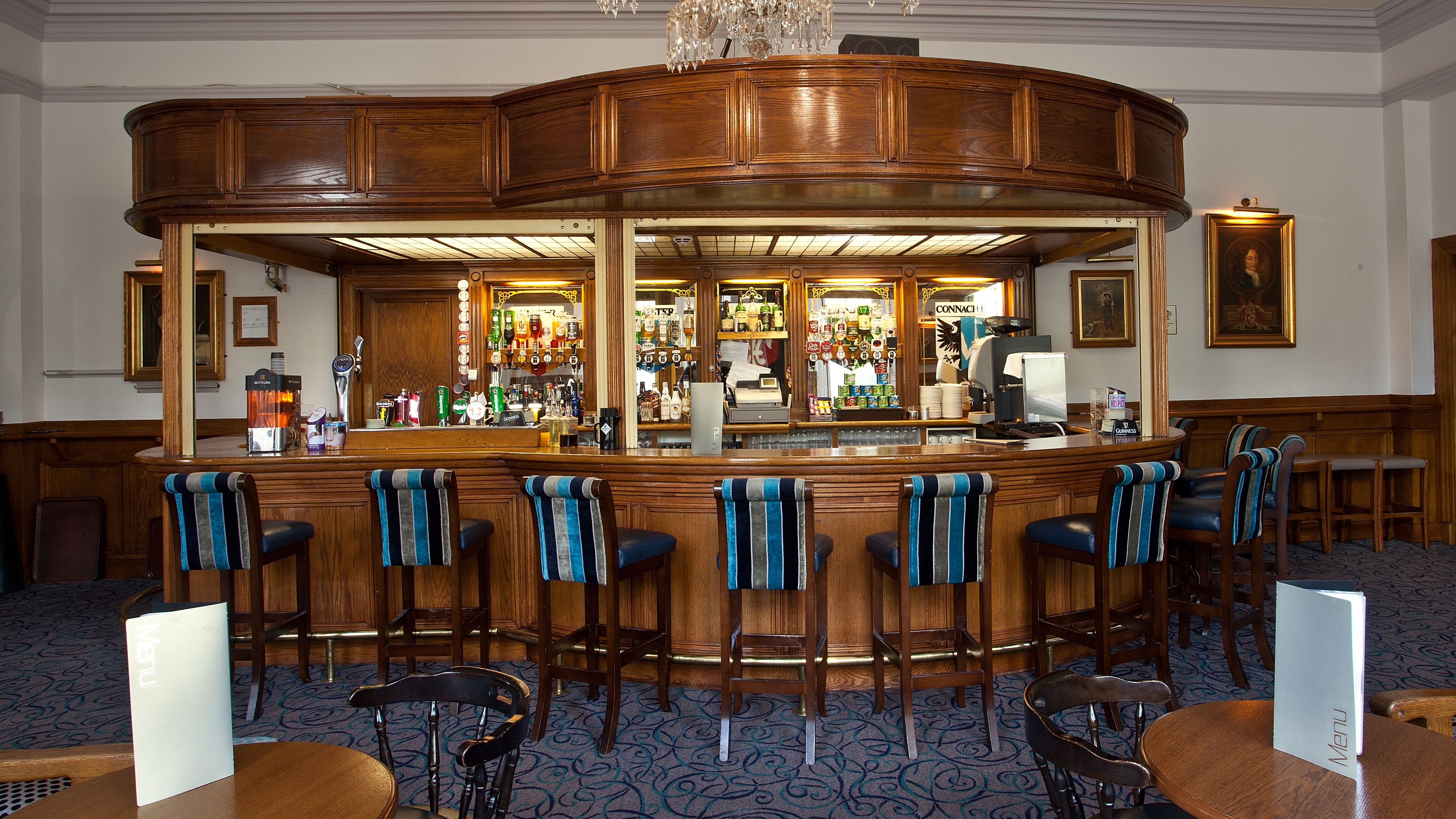 A bar area with a curved wooden counter, bar stools with striped towels, and a variety of bottles and glasses on shelves behind the counter.