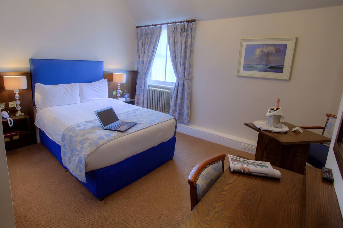 A hotel room with a bed, a desk, a chair, a lamp, a picture on the wall, and a window with curtains.
