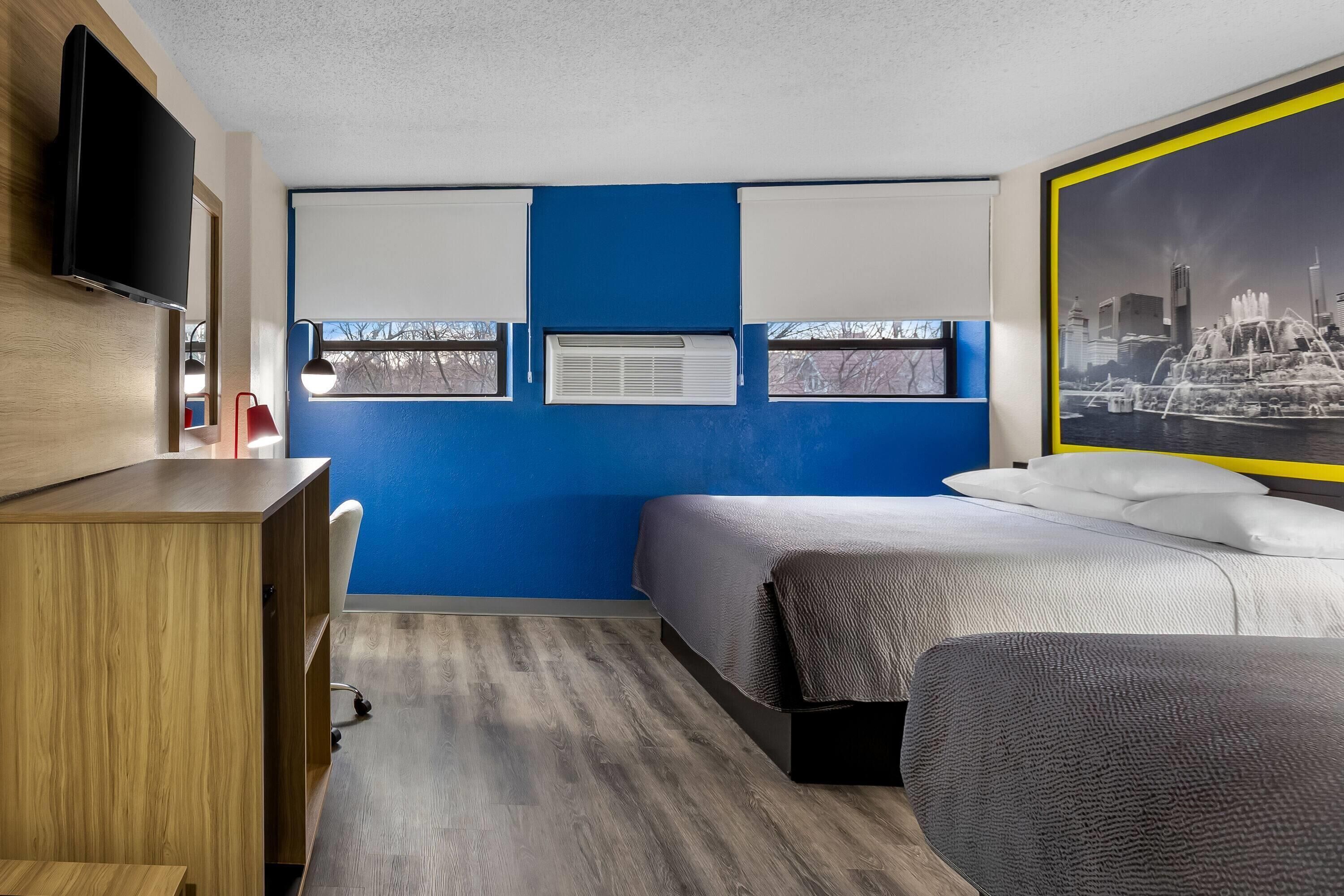 A modern bedroom with a blue accent wall, a bed with a textured gray cover, a wooden desk, a flat-screen TV, and a cityscape poster.