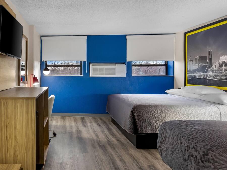 Quarto moderno com parede azul, cama com cobertura cinza texturizada, escrivaninha de madeira, TV de tela plana e pôster com vista para a cidade.