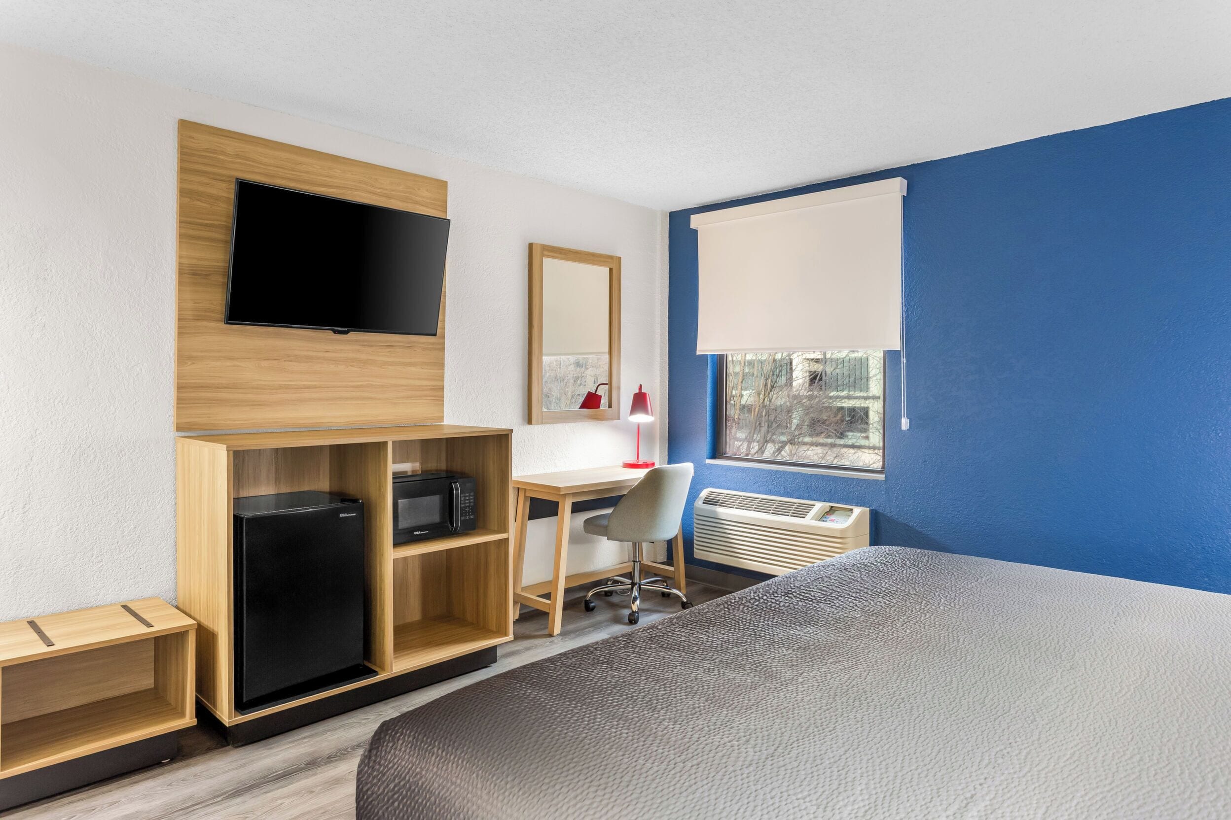 Une chambre d’hôtel comprenant un lit, un bureau avec une chaise, une télévision, un miroir et une fenêtre avec des stores.