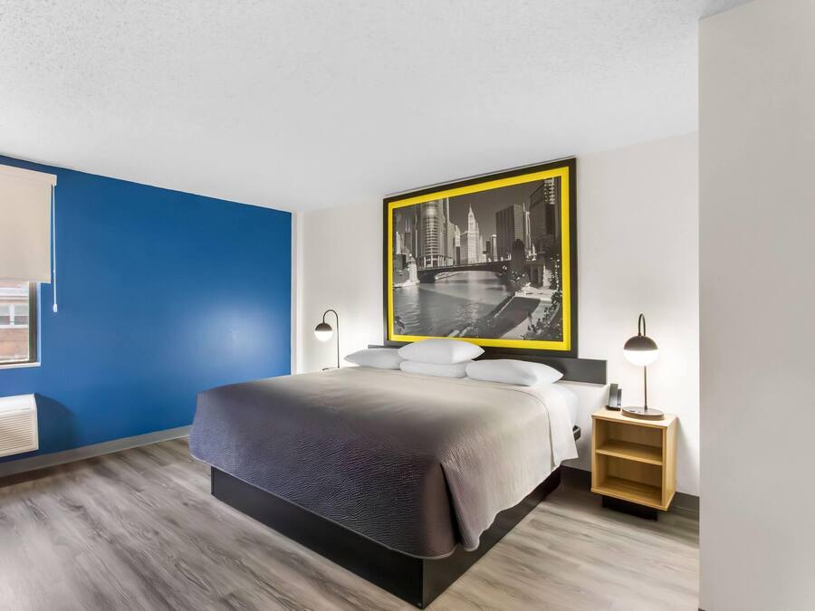 A modern hotel room with a bed, bedside lamps, a nightstand, and a large cityscape painting on the wall.