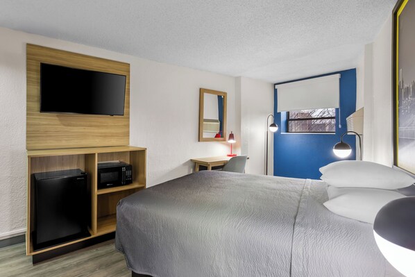 A hotel room with a bed, a desk, a TV mounted on a wooden panel, a microwave, a small refrigerator, a mirror, a lamp, and a window with a view.