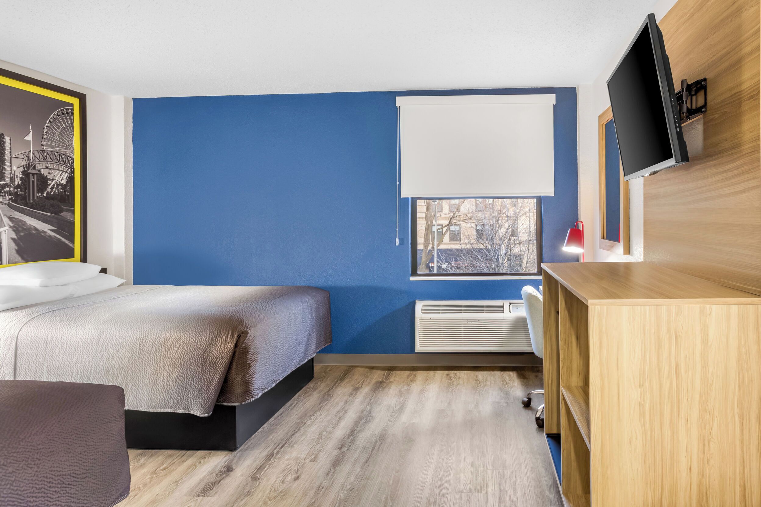 A modern hotel room with a bed, a desk, a TV, and a window with a view of the city.