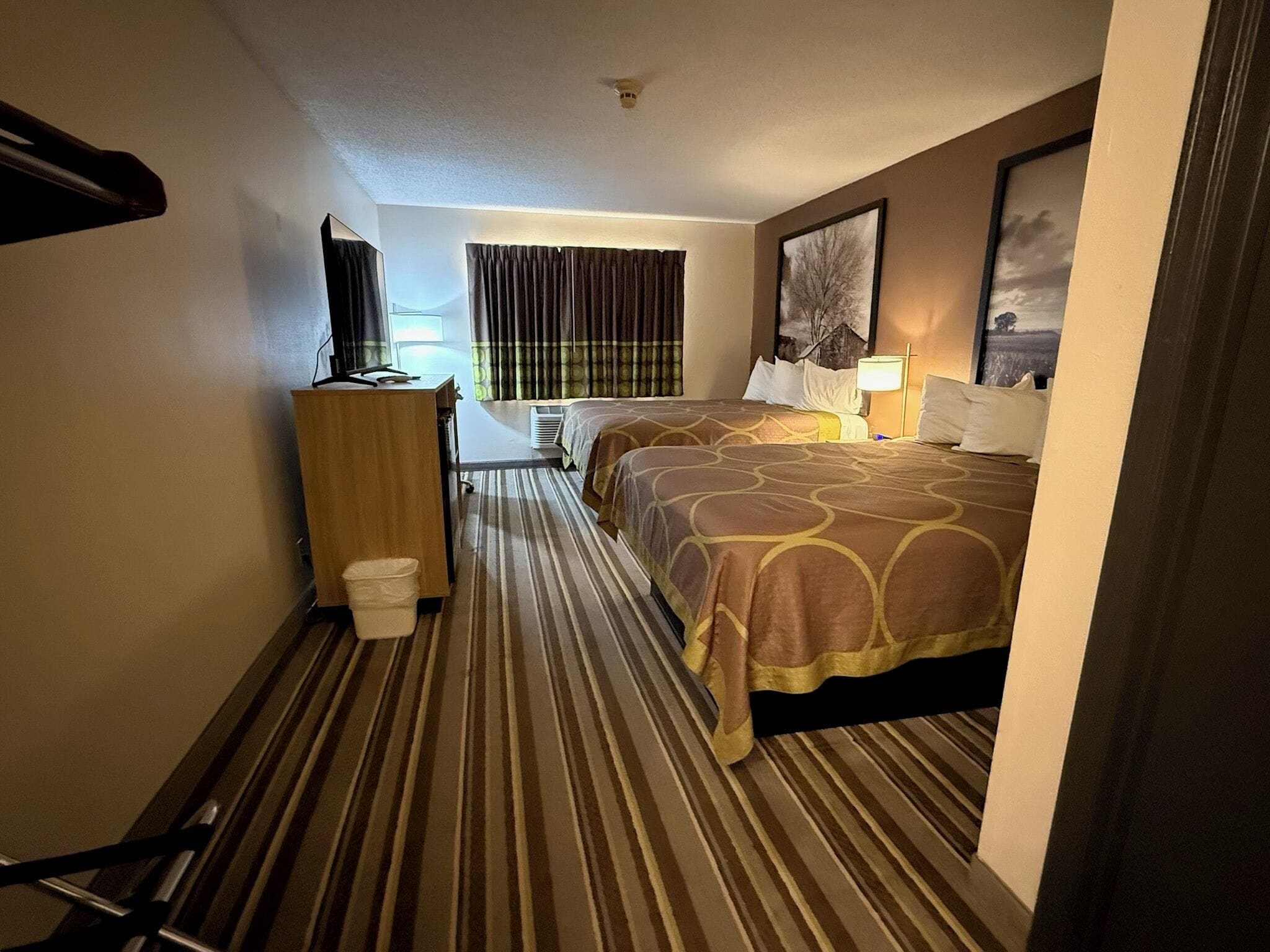 A hotel room with a bed, a desk, a chair, a trash bin, and a window with curtains.