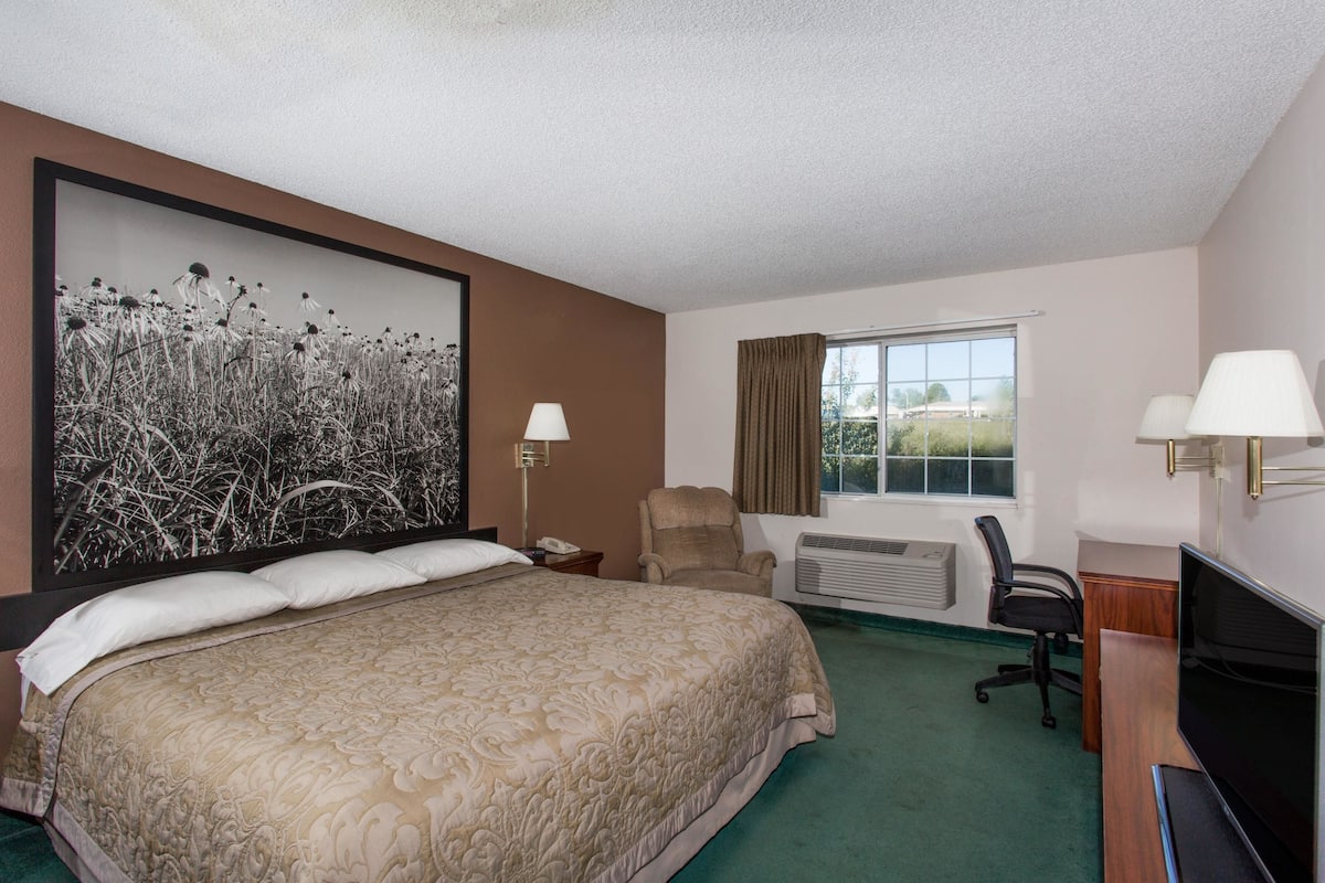 A hotel room with a large bed, a desk with a computer, a chair, a window with curtains, and a wall-mounted TV.