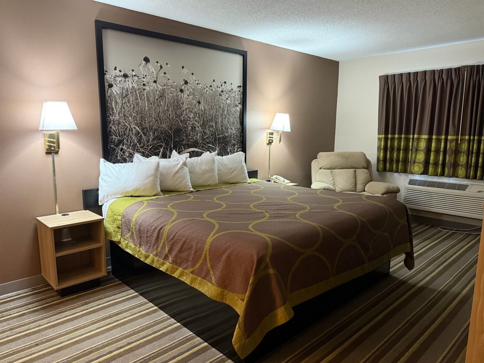 A hotel room with a large bed, two bedside lamps, a chair, a wall-mounted air conditioner, and a large framed picture above the bed.
