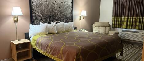 A hotel room with a large bed, two bedside lamps, a chair, a wall-mounted air conditioner, and a large framed picture above the bed.