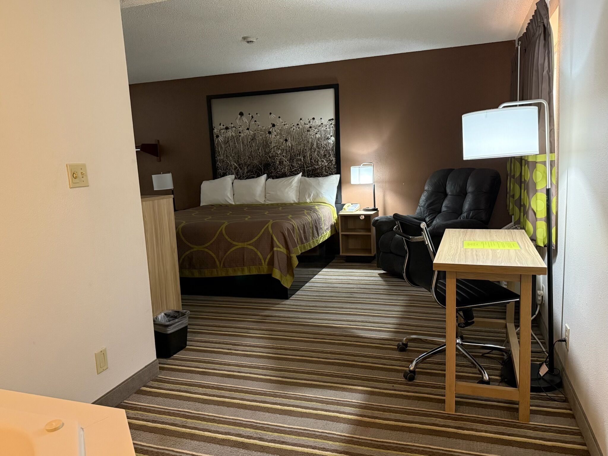 Quarto de hotel com cama, escrivaninha, cadeira, abajur e quadro na parede.