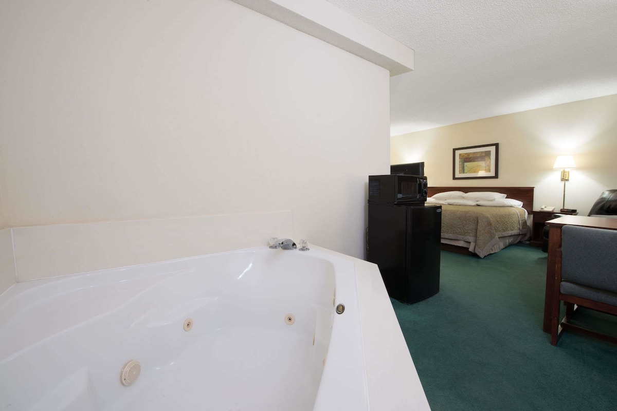 A hotel room with a large white bathtub, a bed with a green blanket, a desk with a chair, and a small black refrigerator.