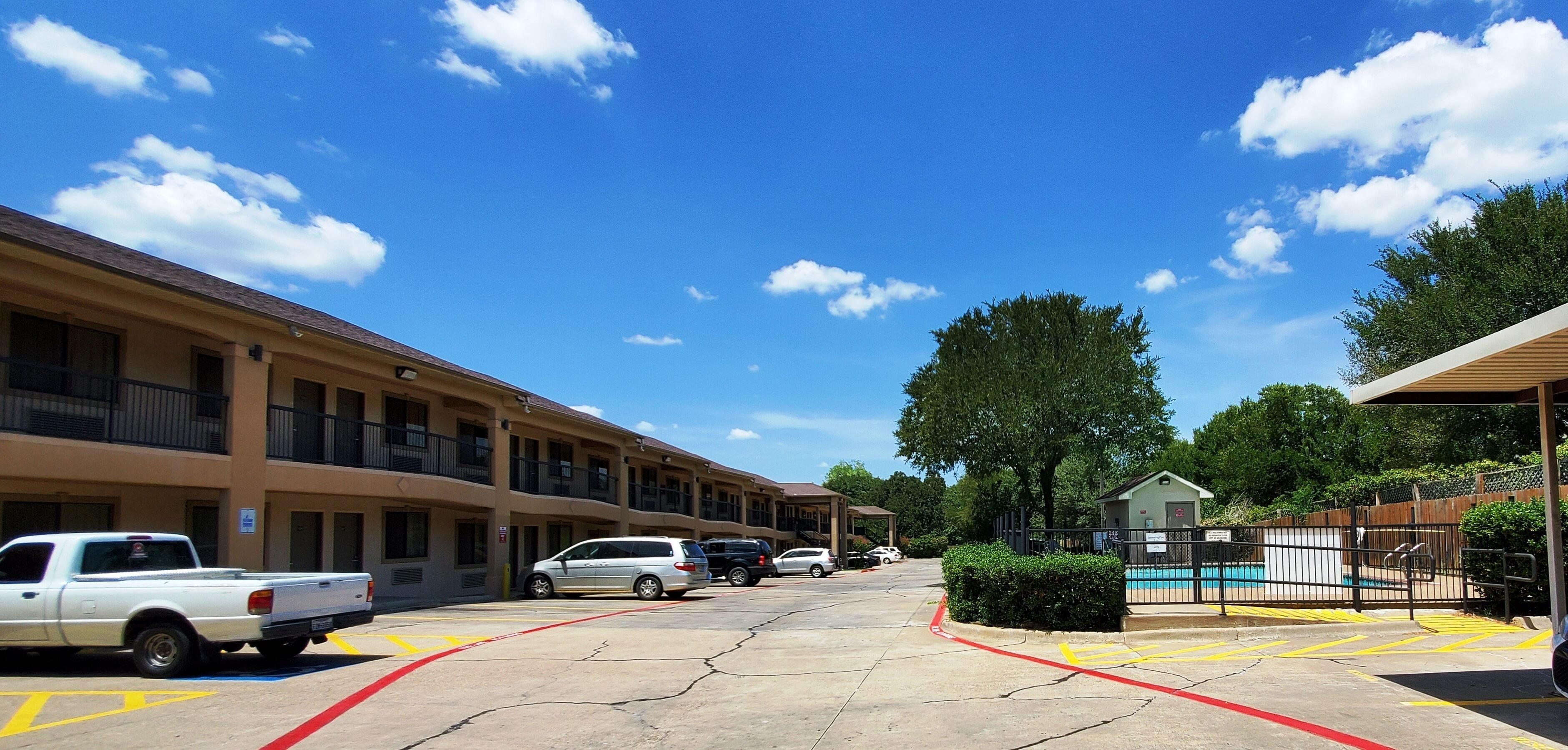 A parking lot with a row of buildings, a swimming pool, and a fenced area.